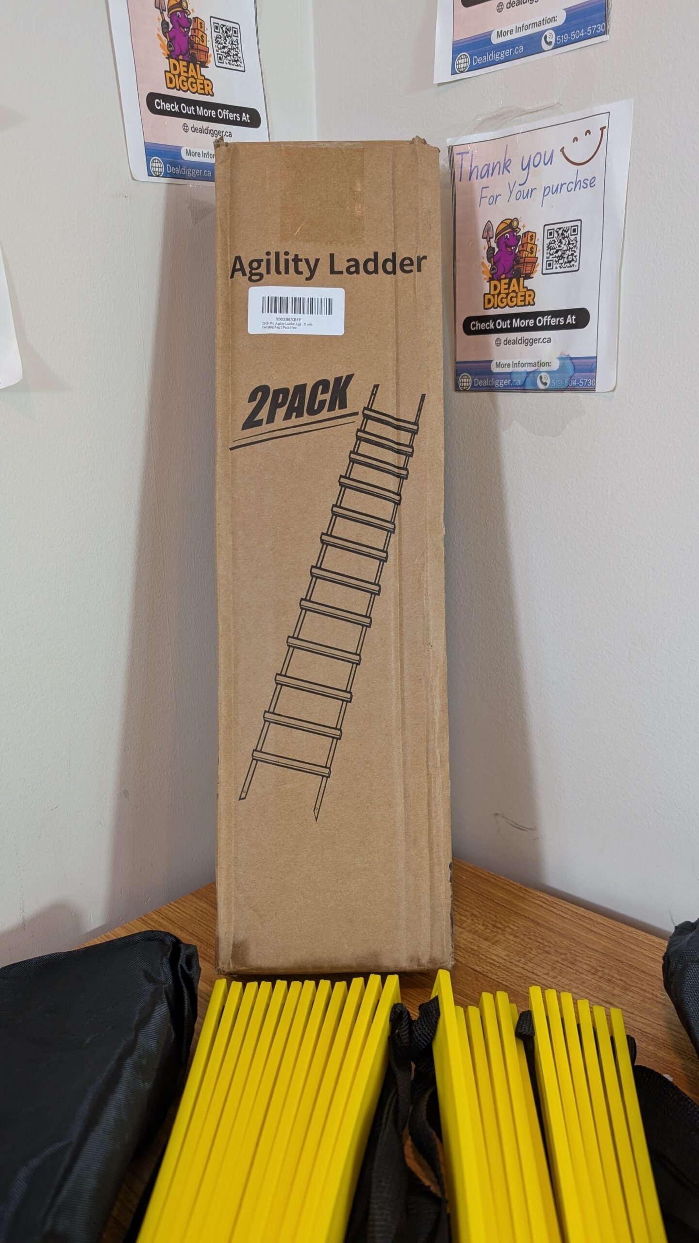 a055ba73-da9e-459f-b717-42b3d1a1e0fd Brown cardboard box for GHB Agility Ladder 2PACK displayed with two sets of yellow ladder rungs and black carrying bags on a wooden surface.